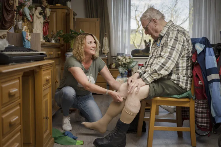 Doelgroep ouderen, begeleider helpt kousen aantrekken bij cliënt die op een stoel zit