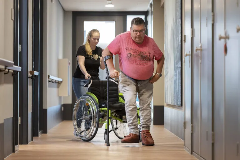 Doelgroep NAH, cliënt stapt uit rolstoel en loopt met kruk door de gang. Zorgmedewerker houdt de rolstoel vast